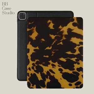 Peut inclure: Une coque pour iPad avec un motif écaille de tortue dans des tons marron et jaune. L'étui est présenté ouvert, révélant un intérieur noir. La marque "BB Case Studio" est visible dans le coin supérieur gauche.