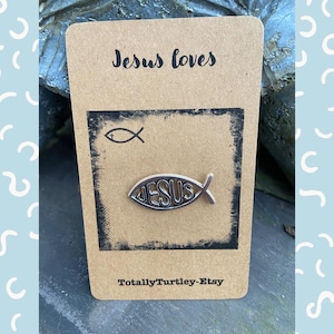 Pu&ograve; includere: Uno spillo a forma di pesce Ges&ugrave; color argento con la parola "JESUS" all'interno del pesce. Lo spillo &egrave; su una carta marrone con il testo "Jesus loves" e "Totally Turtley-Etsy".