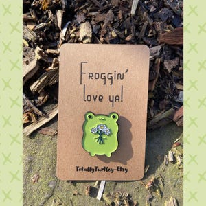 Könnte beinhalten: Ein grüner, froschförmiger Emaille-Anstecker, der einen Strauß weißer Blumen hält, auf einer braunen Karte mit dem Text "Froggin' love ya!". Der Anstecker ist von Totally Turtley-Etsy.