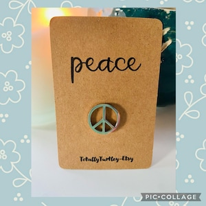 May include: A brown card featuring the word "peace" in black script and a peace symbol pin. The pin is light green with pink and purple hues. The Etsy shop name "TotallyTurtle-Etsy" is printed at the bottom of the card.