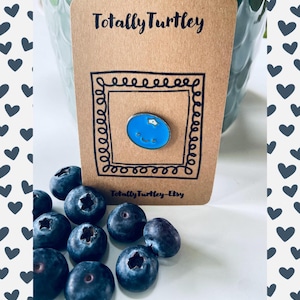 May include: A brown card with the text "Totally Turtley" and a blue enamel pin of a blueberry with a smiling face. The pin is inside a black square frame. Fresh blueberries are scattered around the card.
