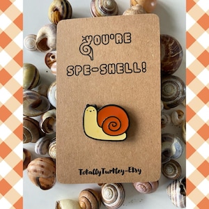 May include: A brown card with the text "You're Spe-Shell!" and a cartoon snail pin with a yellow body and orange shell. The card is surrounded by various seashells. The Etsy shop name "Totally Turtle-Etsy" is printed on the card.