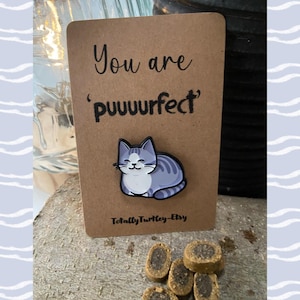 May include: A brown card with the text "You are 'puuurfect'" and a cat pin. The pin is a gray and white cat. The card is on a wooden surface with cat treats in front of it. The Etsy shop name is "TotallyTurtle-Etsy".