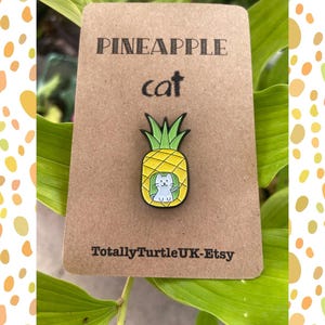 May include: A cat-shaped enamel pin inside a yellow pineapple. The pin is on a brown card with the words "PINEAPPLE cat" and "TotallyTurtleUK-Etsy". The background is green leaves and a yellow and orange dotted border.