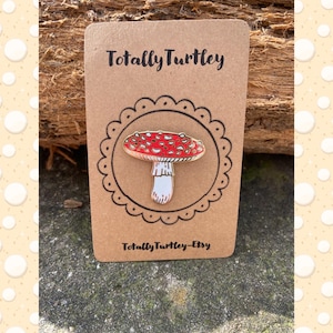 May include: A red and white enamel pin of a mushroom with white spots on a brown card with the text "Totally Turtley" and "Totally Turtley-Etsy".