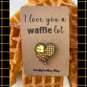 Könnte beinhalten: Eine herzförmige Emaille-Anstecknadel, die eine goldbraune Waffel mit einem schmelzenden gelben Butterstück und Siruptropfen zeigt. Die Anstecknadel ist auf einer braunen rechteckigen Karte mit dem Text I love you a waffle lot und TotallyTurtley-Etsy befestigt.