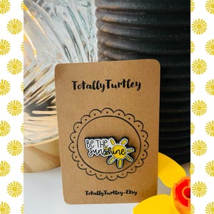 Könnte beinhalten: Eine braune Karte mit dem Text "Totally Turtley" und einer Anstecknadel mit der Aufschrift "Be the Sunshine". Die Anstecknadel ist ein gelbes und weißes Sonnendesign. Die Karte befindet sich auf einem weißen Hintergrund mit gelben Sonnen.