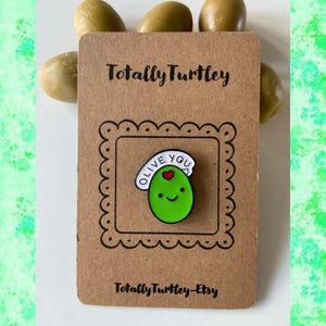 Könnte beinhalten: Eine braune Karte mit dem Text "Totally Turley" und "Totally Turley-Etsy" zeigt eine grüne Oliven-Anstecknadel mit einer Kochmütze mit der Aufschrift "Olive You". Mehrere grüne Oliven sind über der Karte platziert.