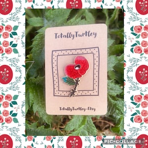 May include: A red poppy brooch with a black stem and green leaves is shown on a brown card. The card features the text "Totally Turtley" and "Totally Turtley-Etsy". The poppy has a glittery texture and is set against green foliage.