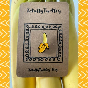 Könnte beinhalten: Ein gelb-oranger Bananen-Emaille-Pin auf einer braunen Karte mit dem Text "Totally Turtley" und "Totally Turtley-Etsy". Der Pin befindet sich in einem schwarzen quadratischen Rahmen mit einem lockigen Design. Der Hintergrund zeigt gelbe Bananen.