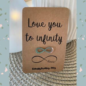 Könnte beinhalten: Eine braune Karte mit den Worten "Love you to infinity" in schwarzer Schrift. Eine Unendlichkeits-Symbol-Anstecknadel mit grünen und rosa Akzenten ist angebracht. Das Wort "love" steht unter einem schwarzen Unendlichkeitszeichen.