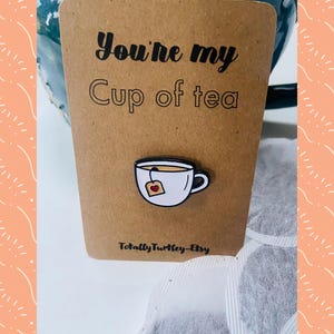 Puede incluir: Una tarjeta marrón con el texto "You're my Cup of tea" y un pequeño pin de esmalte de una taza de té. El pin de la taza de té es blanco con un contorno negro, lleno de té y una bolsita de té. El nombre de la tienda Etsy "TotallyTurtle-Etsy" está en la parte inferior.