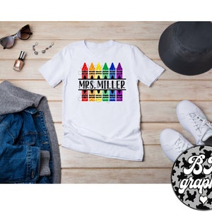 Pu&ograve; includere: Maglietta bianca con un design di matite colorate arcobaleno e il testo "MRS. MILLER" stampato su di essa.