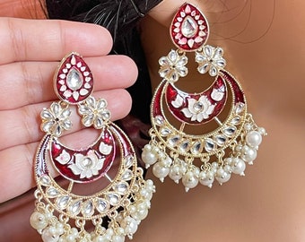 May include: A pair of red and gold earrings with white pearl accents. The earrings feature a teardrop-shaped design with a floral motif and a crescent moon shape.