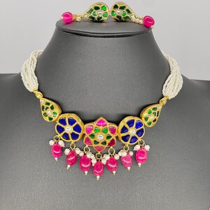 May include: A gold necklace with a multi-colored floral pendant and pink gemstone drops. The necklace is made of white pearls and has a matching pair of earrings with green and pink gemstones.
