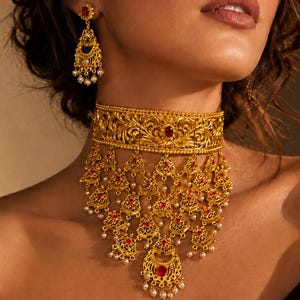 May include: A gold choker necklace featuring intricate filigree metalwork, embedded red stones, and dangling teardrop-shaped pendants accented with small white pearls. Matching drop earrings with a similar gold filigree design and red stone.