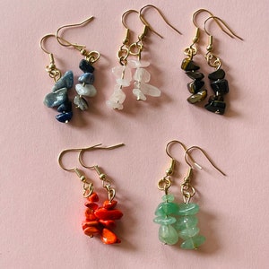 Op de afbeelding: Vijf paar gouden oorhangers met verschillende gekleurde edelsteenchips. De oorhangers zijn verkrijgbaar in verschillende kleuren, waaronder blauw, roze, bruin, oranje en groen.