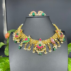 May include: A gold-toned necklace and matching earrings set. The necklace features colorful gemstones in red, green, and white, with dangling pearl accents. The earrings have green gemstone studs and pearl accents. The jewelry is displayed on a black mannequin.