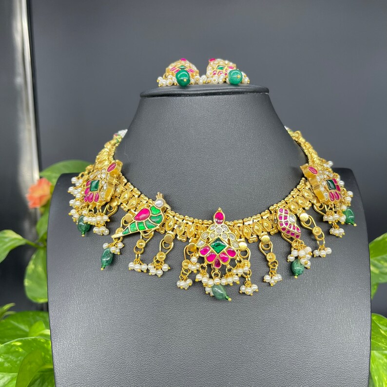 May include: A gold-toned necklace and matching earrings set. The necklace features colorful gemstones in red, green, and white, with dangling pearl accents. The earrings have green gemstone studs and pearl accents. The jewelry is displayed on a black mannequin.
