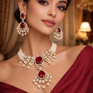 May include: A woman wearing a matching jewelry set featuring a necklace and earrings. The set includes large, square, and teardrop-shaped red gemstones, surrounded by smaller clear stones and white pearls. The jewelry is set against a maroon dress.