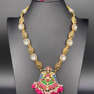May include: A gold necklace with a large, ornate pendant featuring a multi-colored floral design. The pendant is surrounded by pink and white beads. The necklace is on a black display stand.