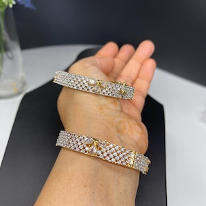 May include: Two gold bangles with a row of clear cubic zirconia stones. The bangles are on a person's wrist.