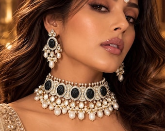 May include: A jewelry set consisting of a choker necklace, drop earrings, and a maang tikka. Each piece features oval black stones framed by small white crystals and accented with dangling white pearls set in a gold-toned metal base.
