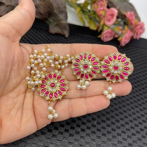 May include: Gold-toned jewelry set featuring a necklace and earrings. The necklace has a floral pendant with a central red stone, surrounded by pink petals and white accents, and a pearl chain. The earrings mirror the pendant design, with a cluster of pearls.