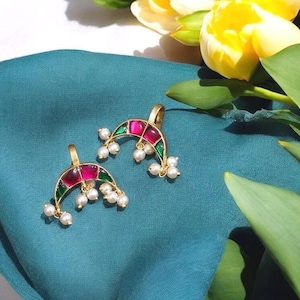 May include: Two gold-coloured earrings with a crescent shape. Each earring has a central red gemstone, green detailing, and small white pearls. They are presented on teal fabric, with yellow tulips visible in the background.