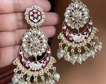 May include: A pair of long, dangling earrings with a red and white enamel design. The earrings feature a large, round, floral-shaped pendant with a crescent moon design. The pendants are adorned with clear stones and pearls.