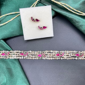 May include: A silver choker necklace with five strands of white pearls and pink accents. The necklace has a traditional design with small, intricate details. The necklace is accompanied by a pair of matching earrings with pink stones.