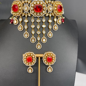 Può includere: Una collana choker dorata con pietre rosse e bianche. La collana ha un design pendente con più file di pietre. Il set include anche un paio di orecchini abbinati.