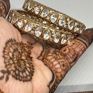 May include: Two gold-toned bangles with clear, oval-shaped stones. The bangles are held in a hand with intricate henna designs. The jewelry is displayed against a neutral background, highlighting the ornate details.