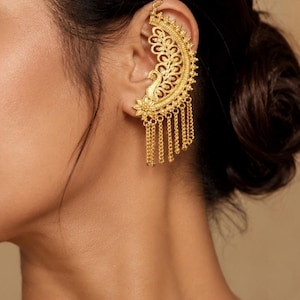 May include: A gold-colored ear cuff featuring an intricate floral and peacock design. The ear cuff dangles with multiple chains. The jewelry is worn on the ear and is a statement piece.