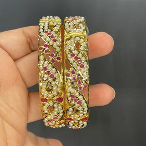 May include: Two gold bangles with intricate designs, adorned with white pearls and pink gemstones.