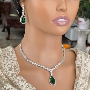May include: A silver necklace with a teardrop-shaped green stone pendant and matching earrings. The necklace is set with small clear stones.