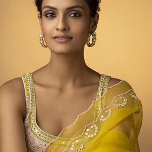 May include: A woman wearing a yellow sari with a matching blouse. The blouse has a white and pink floral design. The sari is draped over the shoulder and has a delicate, embroidered border. The woman is also wearing gold hoop earrings.