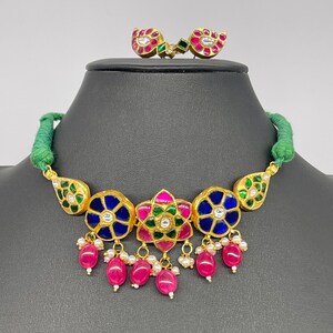 Op de afbeelding: Een goudkleurige ketting en bijpassende oorbellen set. De ketting heeft blauwe en groene bloemmotieven met roze en rode accenten en bungelende roze kralen. De oorbellen hebben een vergelijkbaar ontwerp. De ketting wordt getoond op een zwarte mannequin.