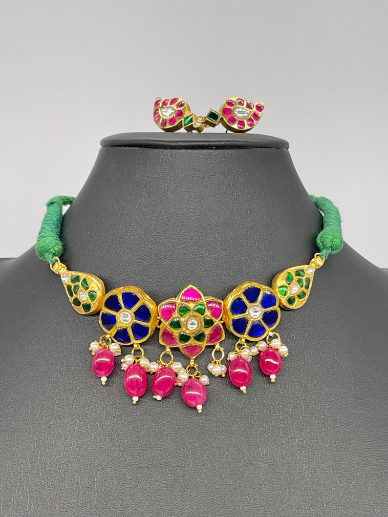 Op de afbeelding: Een goudkleurige ketting en bijpassende oorbellen set. De ketting heeft blauwe en groene bloemmotieven met roze en rode accenten en bungelende roze kralen. De oorbellen hebben een vergelijkbaar ontwerp. De ketting wordt getoond op een zwarte mannequin.
