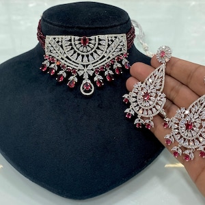 May include: A silver choker necklace with a red stone design and matching earrings. The necklace has a large, round, openwork design with red stones around the edge. The earrings are teardrop shaped with a red stone at the bottom.