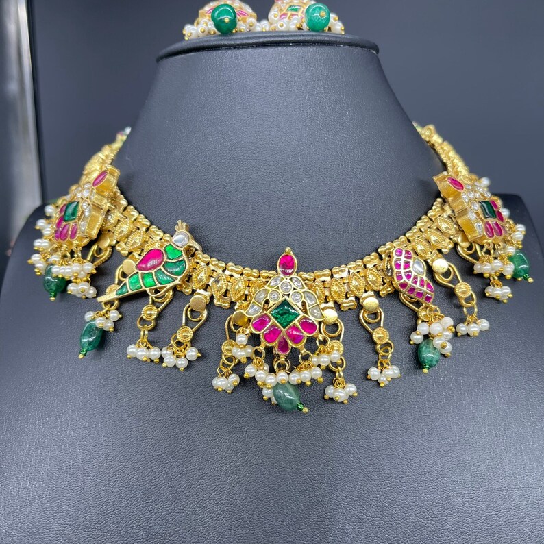 May include: A gold-toned necklace and matching earrings set. The necklace features colorful gemstones, including green, pink, and white, with a bird design. Small white pearls and green beads dangle from the necklace. The earrings have green gemstones.