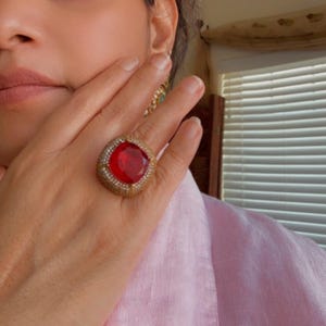 May include: A large, gold-toned ring with a square, faceted, red gemstone. The ring is surrounded by a halo of small, clear stones. The ring is on a finger, and the person is wearing a pink shirt.