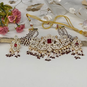 May include: A gold choker necklace with red and white beads, a large ornate pendant with red stones, and matching earrings. The necklace has a layered design with multiple strands of beads.