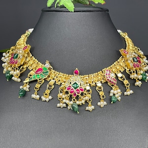 May include: Gold-toned necklace with intricate detailing. Features peacock and floral designs with red, green, and white accents. Embellished with small pearls and green gemstone drops, displayed on a black mannequin.
