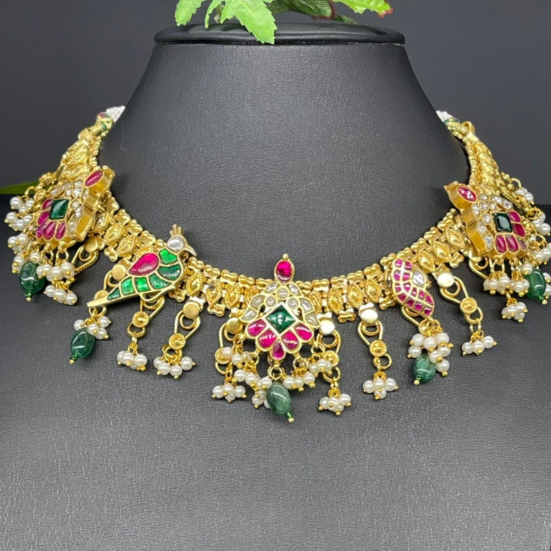 May include: Gold-toned necklace with intricate detailing. Features peacock and floral designs with red, green, and white accents. Embellished with small pearls and green gemstone drops, displayed on a black mannequin.
