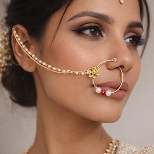 May include: Close-up of gold Indian bridal jewellery. A nose ring with a floral design and pearl and red bead accents is connected to an ear chain. A matching headpiece and necklace are also visible.