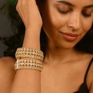 May include: Two gold-colored bangles with clear, round gemstones are worn on a person's wrist. The bangles have a repeating pattern of gemstones set in a gold metal frame. The person is wearing a black top.
