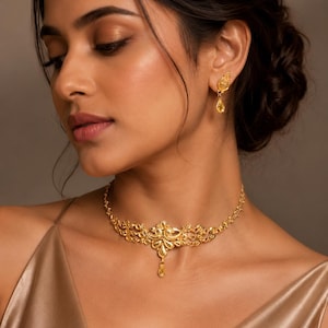 Op de afbeelding: Een goudkleurige ketting en bijpassende oorbellen set. De ketting heeft een sierlijk bloemmotief met een centrale hanger en een kleine, hangende edelsteen. De oorbellen hebben een vergelijkbaar ontwerp met een hangende edelsteen.