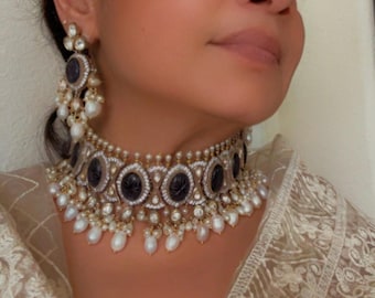 May include: An elaborate necklace and earring set. The choker necklace has dark oval stones, encircled by small clear stones and pearls. The earrings echo the design, with dark stones and pearls. The jewellery is set against a neutral background.