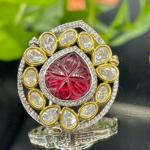 May include: A silver ring with a red gemstone surrounded by clear stones. The ring has a gold band and is set with a carved design.